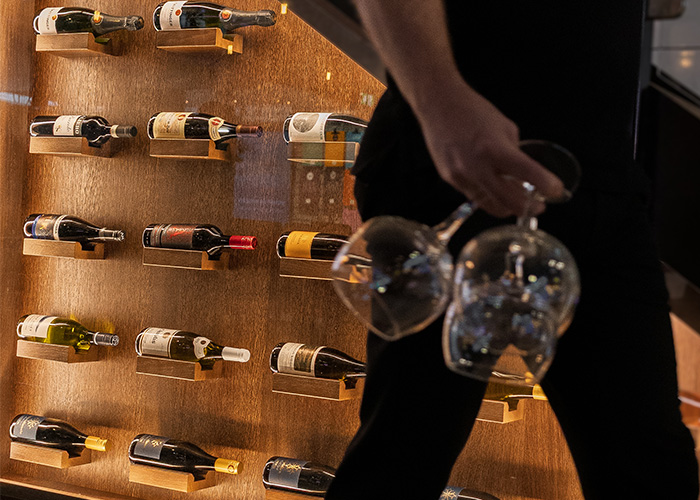Elegant vinvägg med upplysta flaskor i en stämningsfull vinbar, där serveringspersonal passerar med vinglas.